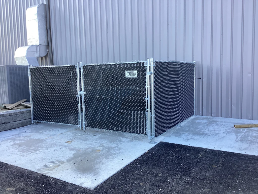 A 6-foot galvanized chain-link fence enclosure with black privacy slats and a double swing gate installed on a concrete pad