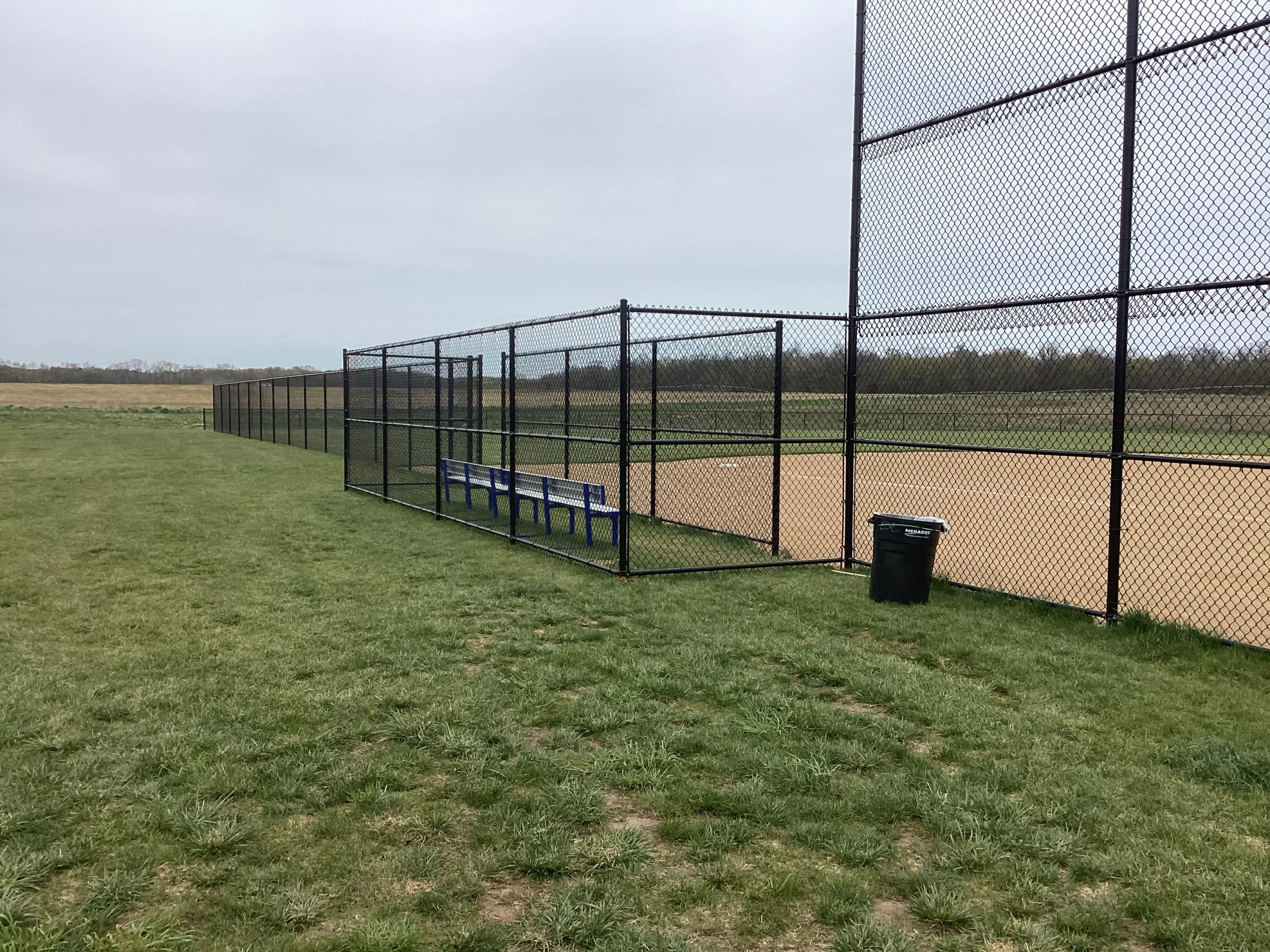A baseball field dugout area enclosed with black vinyl-coated chain-link fencing, including a bench and nearby trash receptacle