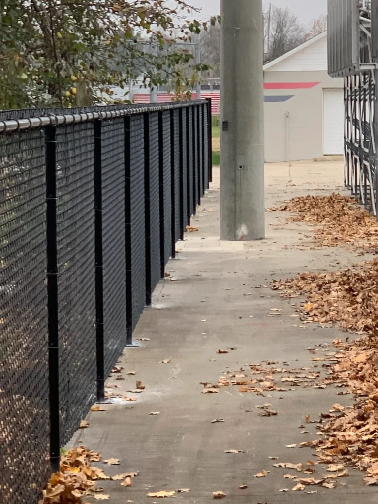A black vinyl-coated chain-link fence bordering a concrete walkway covered in fallen autumn leaves next to a sports stadium structure