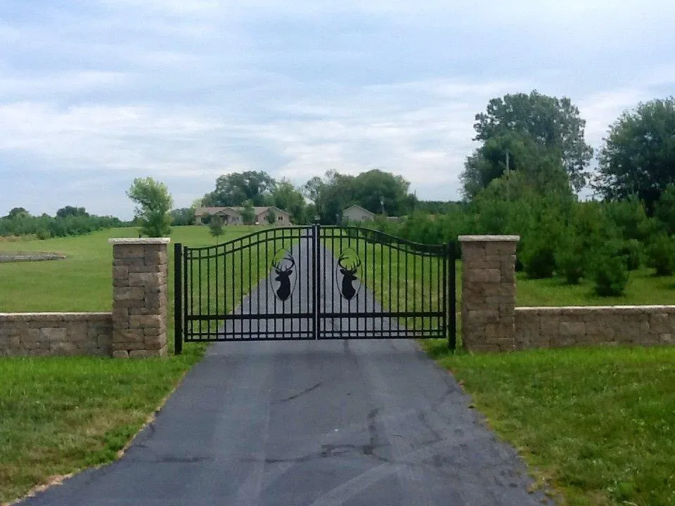 A decorative black arched driveway gate featuring metal deer silhouette inserts, positioned between two stacked-stone columns
