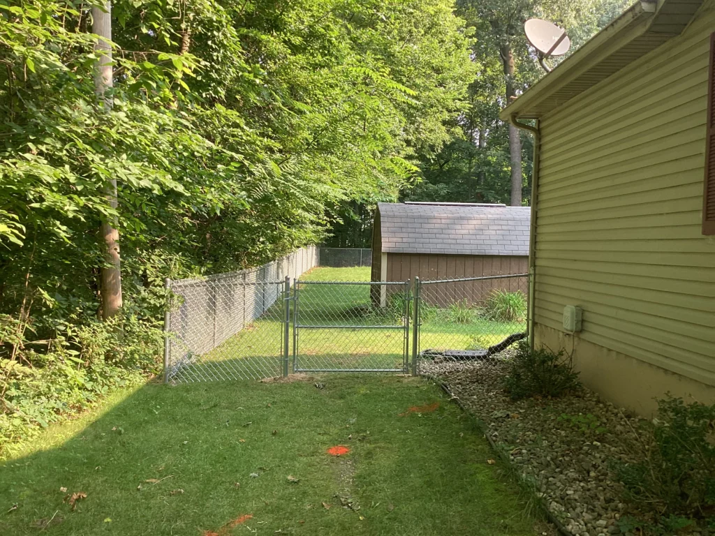 A galvanized chain-link fence and matching gate installed between a residential house with a satellite dish and a dense wooded area