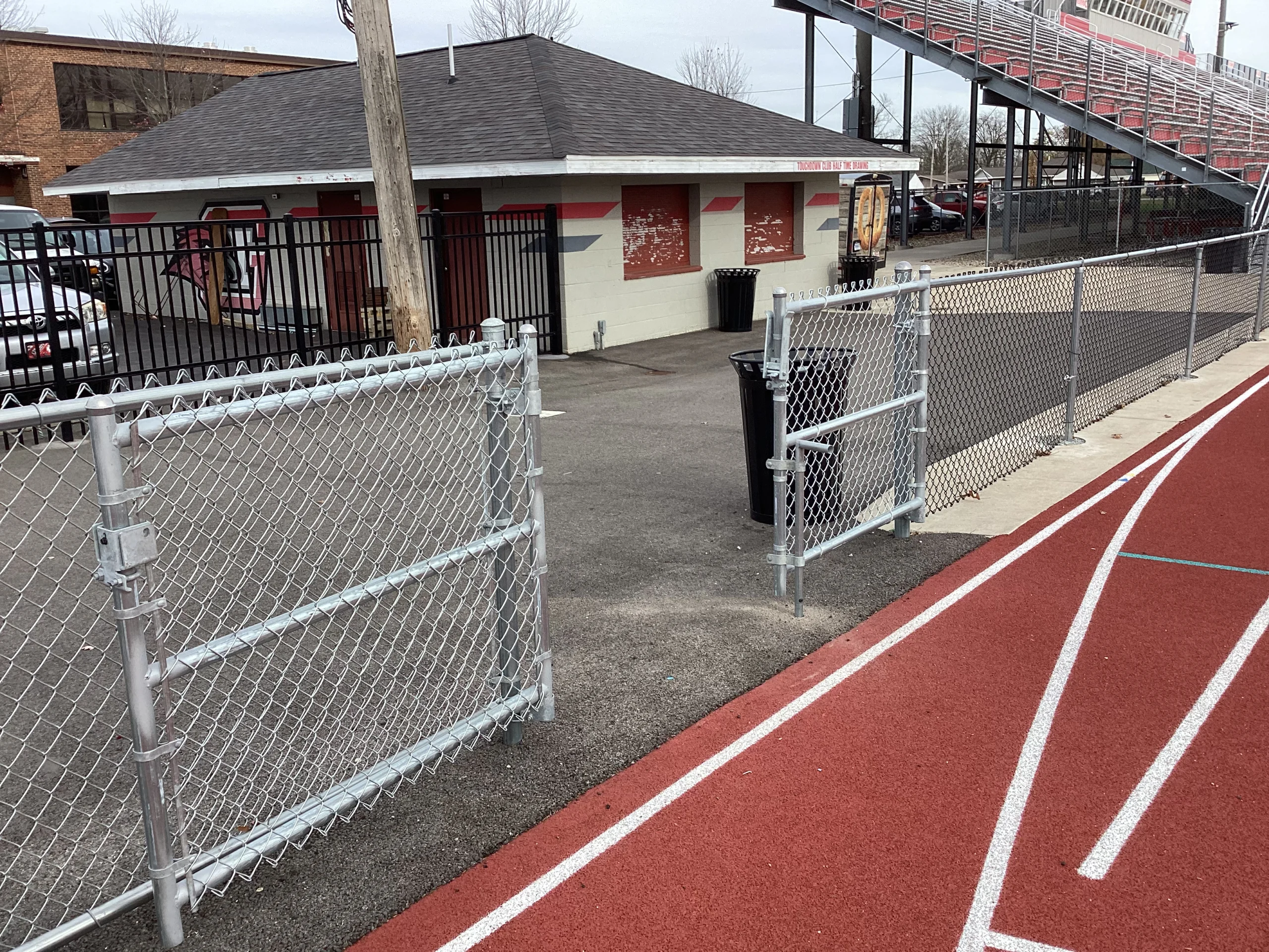 A galvanized chain-link gate and fence transition into a black ornamental fence near a school running track and concession building