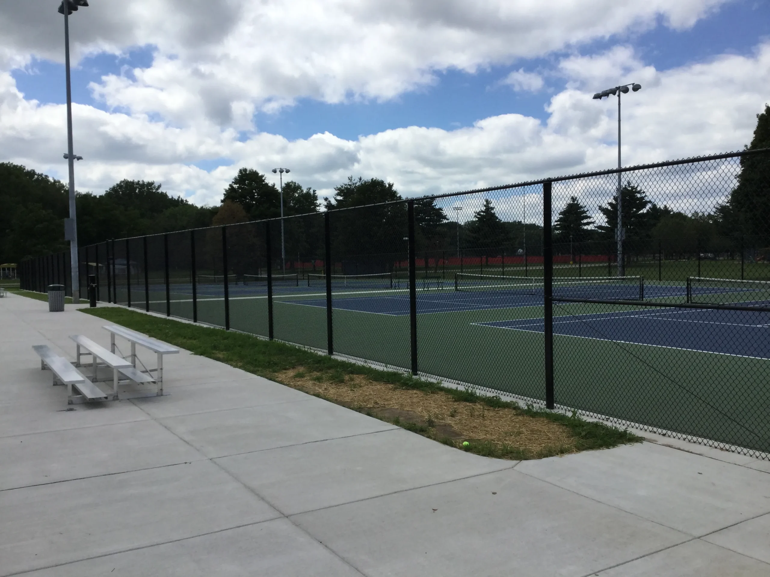 A high-profile black vinyl-coated chain-link fence enclosing multiple blue and green tennis courts next to a concrete spectator area with bleachers