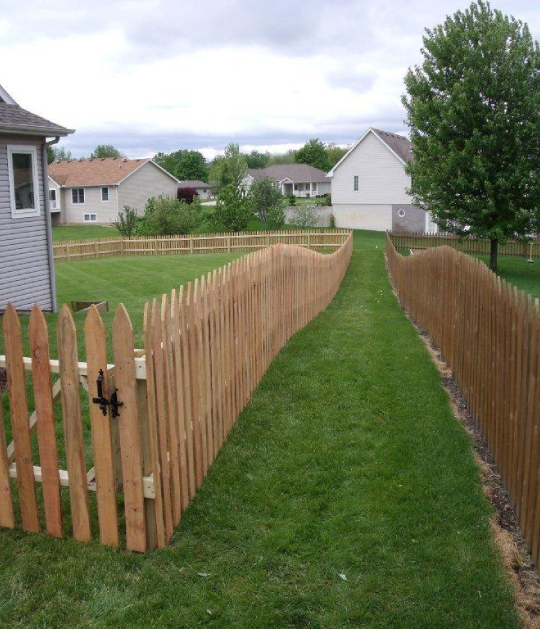 A narrow green grass pathway is enclosed by two wooden picket fences that curve inward, leading to a residential neighborhood with houses and trees.